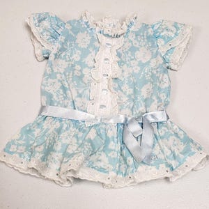 May include: A light blue and white floral print dress featuring short ruffled sleeves, a high neckline with white eyelet lace trim, a vertical center panel of eyelet lace, and a light blue satin ribbon tied at the waist with a flared hem.