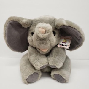 May include: A gray plush elephant toy with large ears, dark eyes, and a light pink trunk tip. The stuffed animal features a tag that reads Soft N Cuddly and AA PLUSH with a small bear graphic. The toy has a soft, fuzzy texture and gray paw pads.