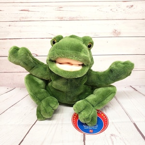 May include: A green plush frog hand puppet with a wide mouth, large dark eyes, and soft textured fabric. The puppet features a round tag with text that reads: It's a frog. Would you put me on your hand? Then put me on your hand. And I'll ribbit for you.