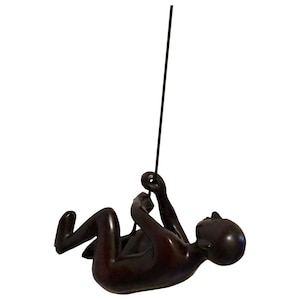 May include: A black metal sculpture of a person hanging from a rope. The sculpture is in a dynamic pose, with the person's arms and legs outstretched. The sculpture is a modern and abstract representation of a climber.