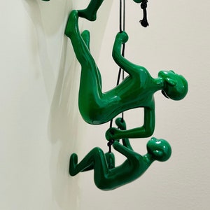 May include: Three glossy green climbing figures on a wall, each suspended by a black rope. The figures are in various climbing poses, creating a dynamic and decorative wall art piece. The figures are attached to a white wall.