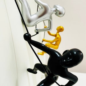 May include: Three metal figurines of people rock climbing, hanging from ropes. The figurines are silver, gold, and black.