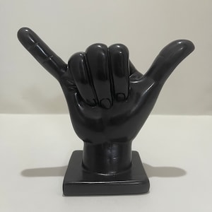 May include: A black sculpture of a hand in the shaka gesture, with the thumb and pinky extended. The hand is mounted on a square base. The sculpture is made of a glossy, solid material.