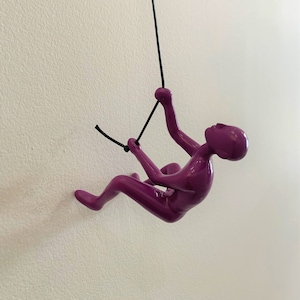 May include: A purple metal figurine of a person rock climbing, hanging from a black rope.