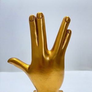 Peut inclure: Une sculpture en forme de main dorée dans la pose du salut vulcain. La sculpture est montée sur une base carrée. Les doigts sont séparés dans le geste emblématique, le pouce étant tendu. La sculpture est en matériau métallique brillant.