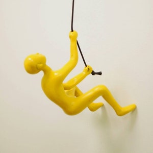 May include: A yellow figurine of a person climbing a rope, hanging from a white wall.