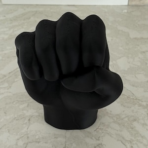 May include: A black sculpture of a clenched fist with a lip-shaped design on the knuckles. The sculpture is made of a solid, matte black material and sits on a marble-patterned surface.