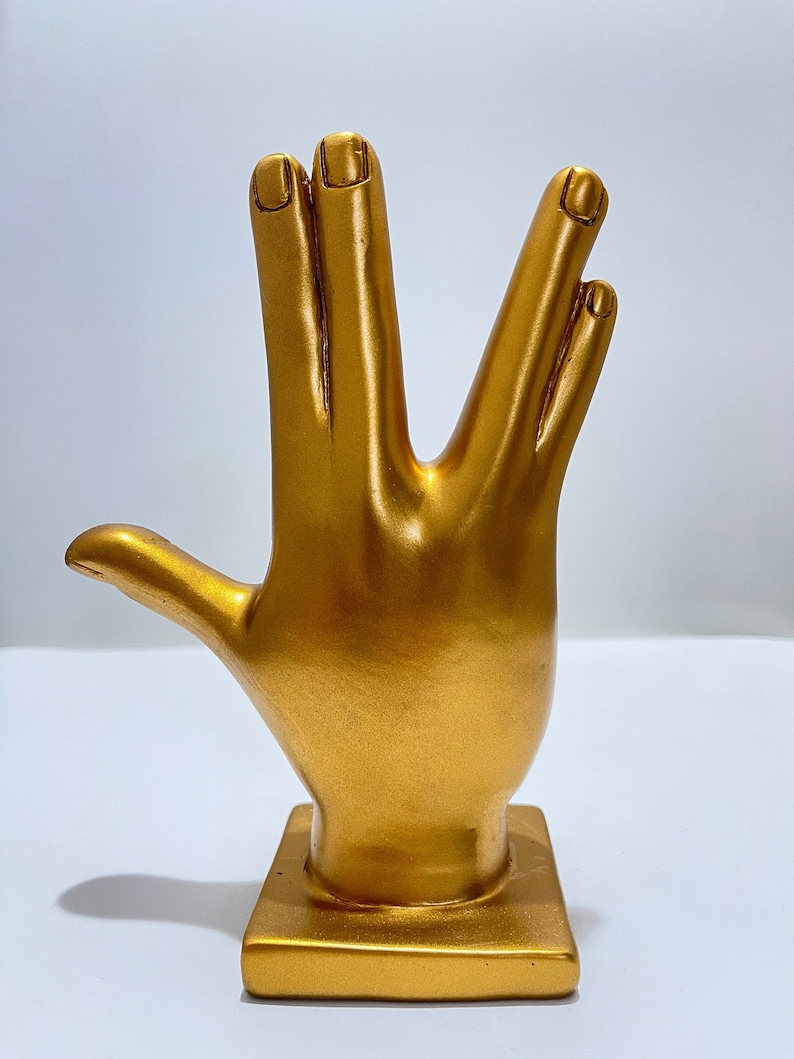 May include: A gold-colored hand sculpture in the Vulcan salute pose. The sculpture is mounted on a square base. The fingers are separated in the iconic gesture, with the thumb extended. The sculpture is made of a shiny, metallic material.