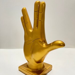May include: A gold-colored hand statue in the Vulcan salute pose. The statue is on a square base and has a smooth, metallic finish. The fingers are spread in the iconic gesture.