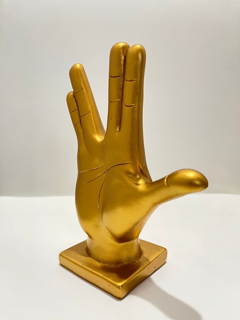 May include: A gold-colored hand statue in the Vulcan salute pose. The statue is on a square base and has a smooth, metallic finish. The fingers are spread in the iconic gesture.