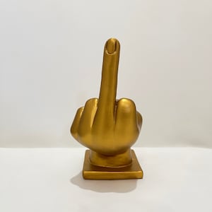 Könnte beinhalten: Eine goldfarbene Handskulptur mit ausgestrecktem Mittelfinger in einer unhöflichen Geste. Die Hand befindet sich auf einem quadratischen Sockel.
