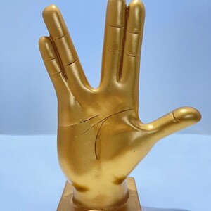 May include: A gold-colored hand statue in the Vulcan salute pose. The sculpture is mounted on a square base and has detailed lines representing the palm and fingers. The statue is a solid color and has a smooth finish.