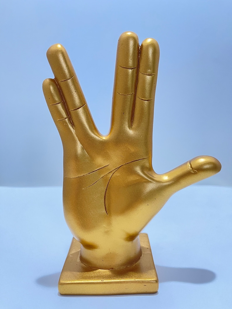 May include: A gold-colored hand statue in the Vulcan salute pose. The sculpture is mounted on a square base and has detailed lines representing the palm and fingers. The statue is a solid color and has a smooth finish.