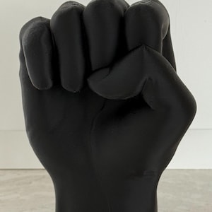 May include: A solid black sculpture of a clenched fist, a symbol of solidarity and resistance. The sculpture is smooth and has a matte finish. The fist is raised, with the knuckles and fingers clearly defined. The base is flat, allowing it to stand upright.