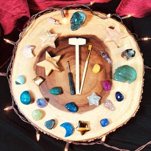 May include: A wooden tray with a variety of crystals, stones, and other objects. The tray is decorated with a light brown wood grain pattern. The objects include a green skull, a hammer, a paintbrush, a star, and a variety of crystals in different colors.