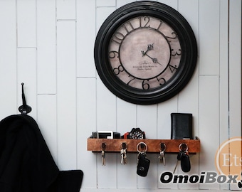 Può includere: Un porta chiavi in legno con ganci e uno scaffale per riporre chiavi, portafogli e altri piccoli oggetti. Il porta chiavi è montato su una parete bianca con un orologio nero sopra.