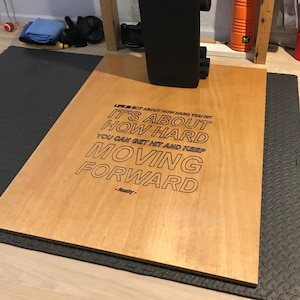 以下が含まれることがあります： 「LIFE IS NOT ABOUT HOW HARD YOU HIT IT'S ABOUT HOW HARD YOU CAN GET HIT AND KEEP MOVING FORWARD -Rooky- 」という励みになる言葉が書かれた木製ウェイトリフティングプラットフォームです。