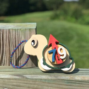 Puede incluir: Una tortuga de madera con una flecha roja y el número 719 en su caparazón. La tortuga está sobre una superficie de madera con un fondo verde.