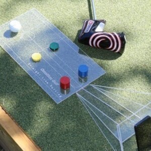 Puede incluir: Un putting green de plástico transparente con una bola de golf blanca y cinco objetivos de colores. Los objetivos son rojo, verde, azul, amarillo y naranja. Una herramienta de putting negra y roja se encuentra en el lado izquierdo de la imagen. Un teléfono inteligente está en un soporte de plástico transparente en el lado derecho de la imagen.