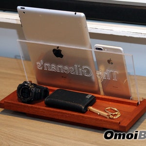 May include: A wooden phone stand with a clear acrylic insert. The stand holds a tablet, a phone, a watch, a wallet, and a key chain. The stand is engraved with the text "The Gilson's".