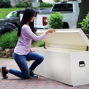 May include: A white storage bench with a hinged lid open. The bench is made of wood and has a black handle. A person is kneeling beside the bench, reaching for the lid.