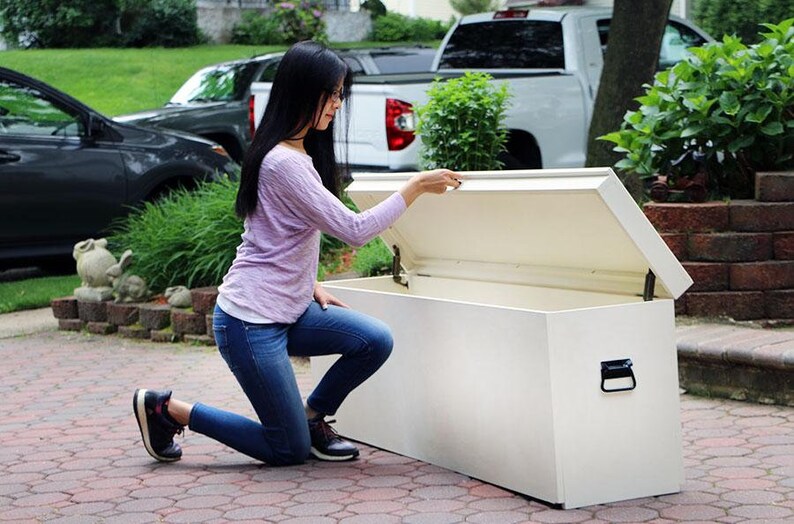 May include: A white storage bench with a hinged lid open. The bench is made of wood and has a black handle. A person is kneeling beside the bench, reaching for the lid.