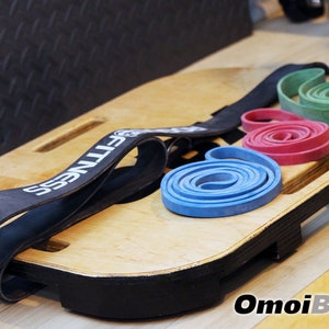 May include: A wooden balance board with a black edge and a black resistance band with the word "FITNESS" printed on it. Three resistance bands are on the board: blue, red, and green.