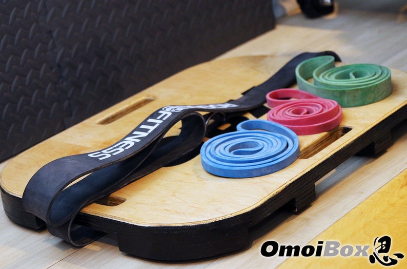 May include: A wooden balance board with a black edge and a black resistance band with the word "FITNESS" printed on it. Three resistance bands are on the board: blue, red, and green.