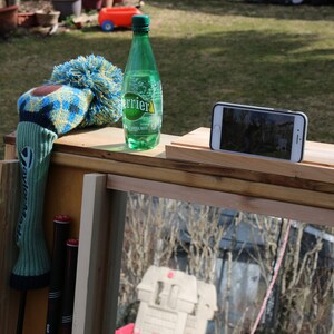 Puede incluir: Un soporte de madera con una botella verde de agua con gas Perrier, una funda para la cabeza de palo de golf azul y amarilla y un tel&eacute;fono inteligente en una peque&ntilde;a plataforma de madera.
