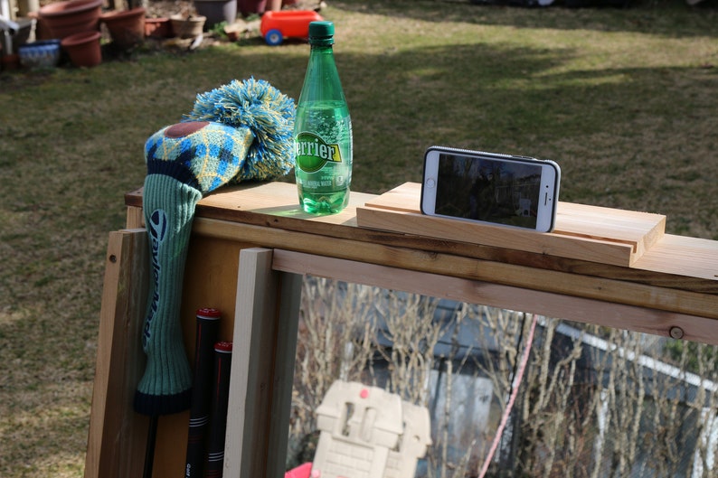 Puede incluir: Un soporte de madera con una botella verde de agua con gas Perrier, una funda para la cabeza de palo de golf azul y amarilla y un tel&eacute;fono inteligente en una peque&ntilde;a plataforma de madera.