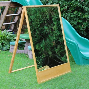 Puede incluir: Un caballete de madera con un espejo sujeto a &eacute;l. El caballete est&aacute; de pie en un c&eacute;sped verde frente a un tobog&aacute;n verde.