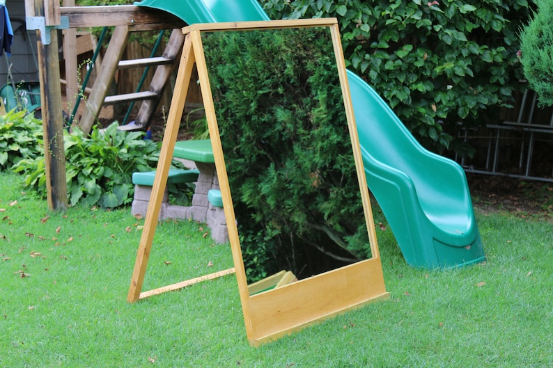 Puede incluir: Un caballete de madera con un espejo sujeto a &eacute;l. El caballete est&aacute; de pie en un c&eacute;sped verde frente a un tobog&aacute;n verde.