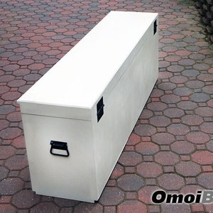 May include: A white wooden storage chest with two black metal handles and two black metal latches. The chest is sitting on a red brick patio.