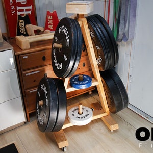 May include: A wooden weight plate storage rack with wheels, holding black and white weight plates with the word "ROGUE" printed on them. The rack is in a home gym setting.