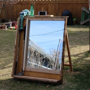 Puede incluir: Un espejo de madera con un estante debajo. El espejo est&aacute; apoyado en un caballete y refleja el cielo y una valla. Hay un palo de golf y una funda de palo de golf verde y azul apoyados contra el espejo.