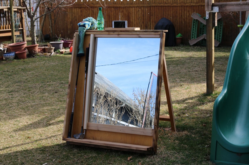 Puede incluir: Un espejo de madera con un estante debajo. El espejo est&aacute; apoyado en un caballete y refleja el cielo y una valla. Hay un palo de golf y una funda de palo de golf verde y azul apoyados contra el espejo.