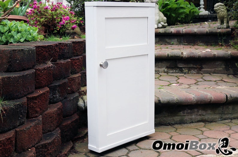 May include: White cabinet with a single door and a silver knob. The cabinet is standing on a brick patio.