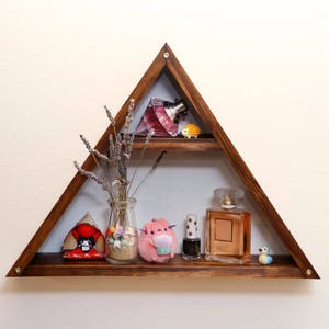 Peut inclure: Une étagère en bois triangulaire à deux niveaux. L'étagère supérieure présente un flacon de parfum rose, une petite figurine jaune et de la lavande séchée. L'étagère inférieure a une petite figurine rouge, un bocal en verre transparent avec de la lavande séchée, un jouet en peluche rose, un flacon de vernis à ongles noir et un flacon de parfum en verre brun.