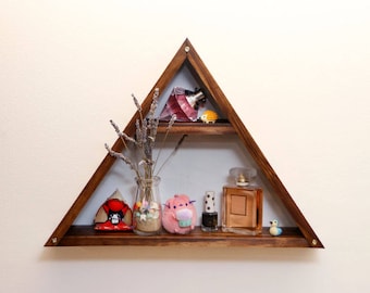 Puede incluir: Una estantería de madera triangular con dos estantes. El estante superior tiene una botella de perfume rosa, una pequeña figurita amarilla y lavanda seca. El estante inferior tiene una pequeña figurita roja, un frasco de vidrio transparente con lavanda seca, un juguete de peluche rosa, una botella de esmalte de uñas negro y una botella de perfume de vidrio marrón.