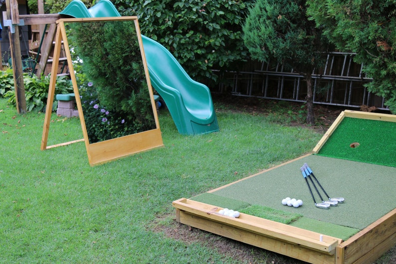 Puede incluir: Un campo de minigolf instalado en un jard&iacute;n con una superficie de putting verde, un marco de madera y un agujero. Hay palos de golf y pelotas de golf en la superficie verde. Un tobog&aacute;n de pl&aacute;stico verde y un espejo de madera est&aacute;n en el fondo.