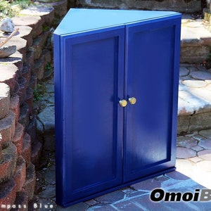May include: A blue corner cabinet with two doors and gold knobs. The cabinet is sitting on a brick patio.