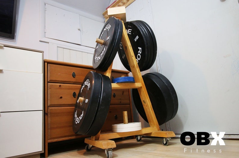 May include: A wooden weight plate storage rack with wheels. The rack is shaped like a triangle and has four shelves. The rack is holding a variety of black weight plates. The rack is labeled "OBX Fitness".