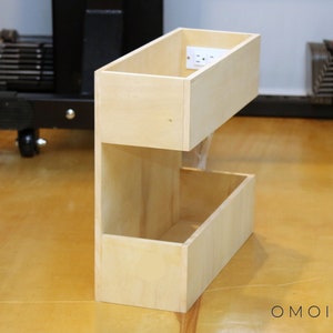 May include: A two-tiered wooden shelf unit with a power outlet in the top shelf. The shelf is made of light wood and has a simple design.