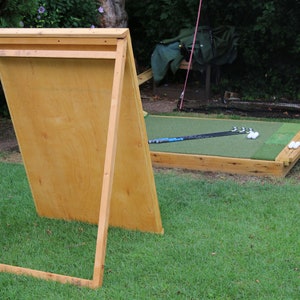 Puede incluir: Una red de pr&aacute;ctica de golf de madera con un tablero trasero de madera contrachapada marr&oacute;n y un &aacute;rea de golpeo de c&eacute;sped artificial verde. Hay palos de golf y pelotas de golf en el c&eacute;sped.