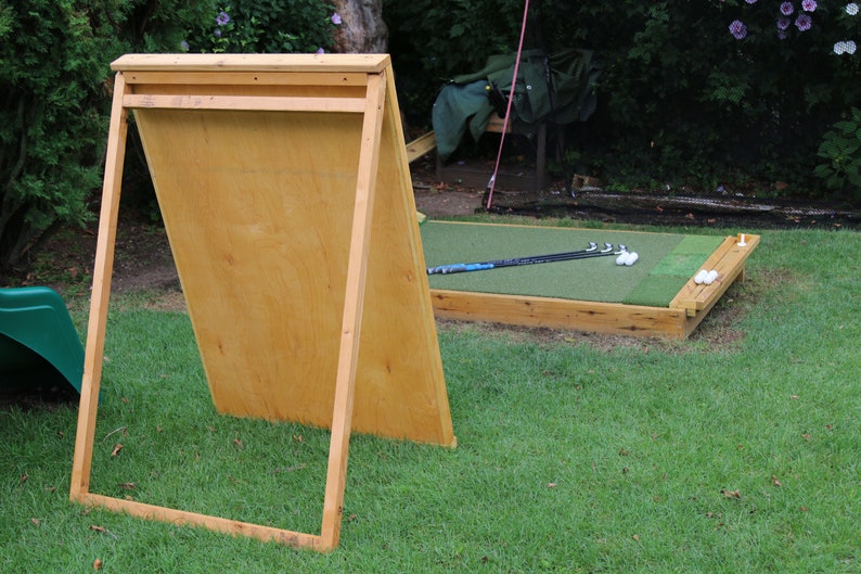 Puede incluir: Una red de pr&aacute;ctica de golf de madera con un tablero trasero de madera contrachapada marr&oacute;n y un &aacute;rea de golpeo de c&eacute;sped artificial verde. Hay palos de golf y pelotas de golf en el c&eacute;sped.