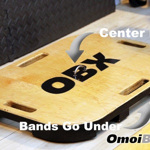 May include: A wooden platform with a black "OBX" logo and a metal hook in the center. The text "Bands Go Under" is written below the platform. The platform is designed for resistance band exercises.