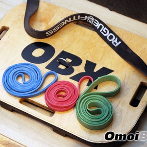 May include: A wooden balance board with the text "OBX" printed on it. The board has a black strap with the text "Rogue Fitness" printed on it. There are three resistance bands on the board: blue, red, and green.