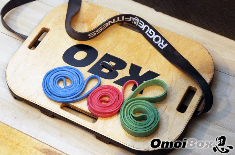 May include: A wooden balance board with the text "OBX" printed on it. The board has a black strap with the text "Rogue Fitness" printed on it. There are three resistance bands on the board: blue, red, and green.