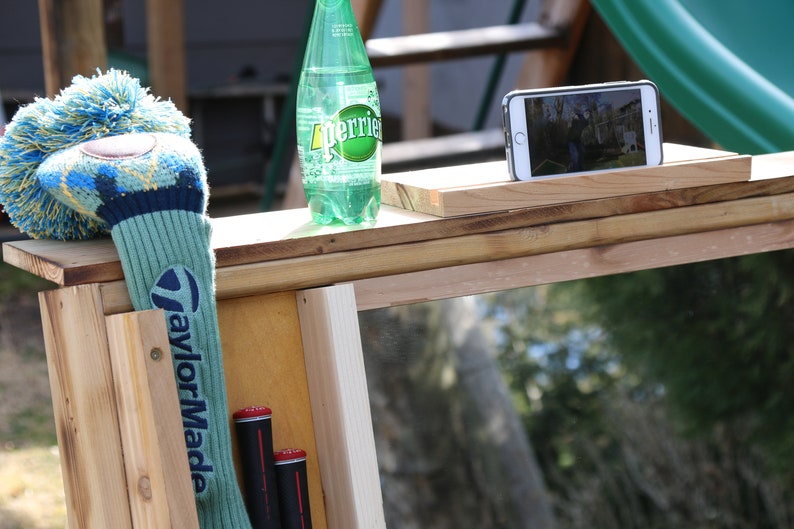 Puede incluir: Una estaci&oacute;n de pr&aacute;ctica de golf de madera con un espejo, una funda para la cabeza de un palo de golf, dos palos de golf y una botella de agua con gas Perrier. La estaci&oacute;n tiene un soporte para tel&eacute;fono en la parte superior.