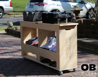 Puede incluir: Un carro de madera con ruedas, etiquetado como "OBX Fitness", contiene un juego de mancuernas ajustables, un par de zapatillas blancas y otros equipos de fitness.
